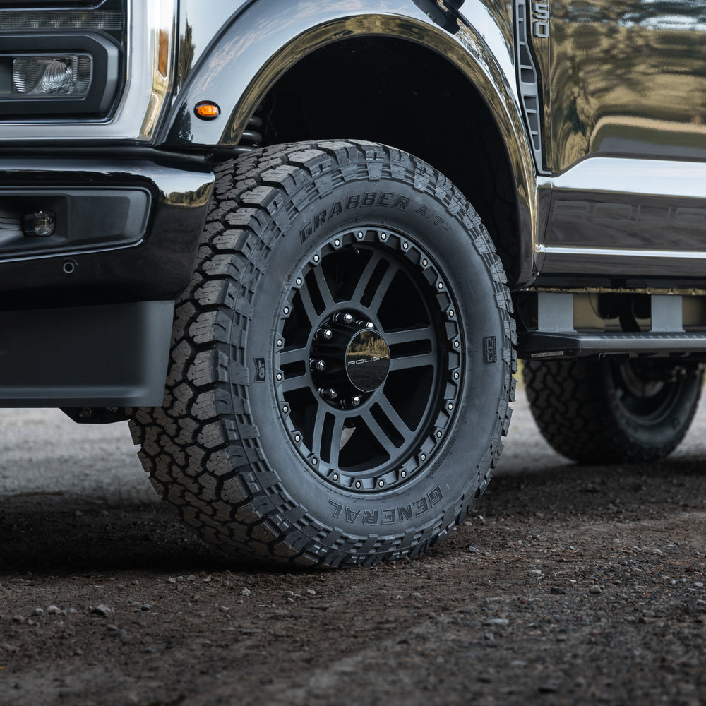 
                  
                    Close-up of a black Roush Super Duty truck tire and wheel parked on a gravel surface, showing the tread and part of the vehicle's front side.
                  
                