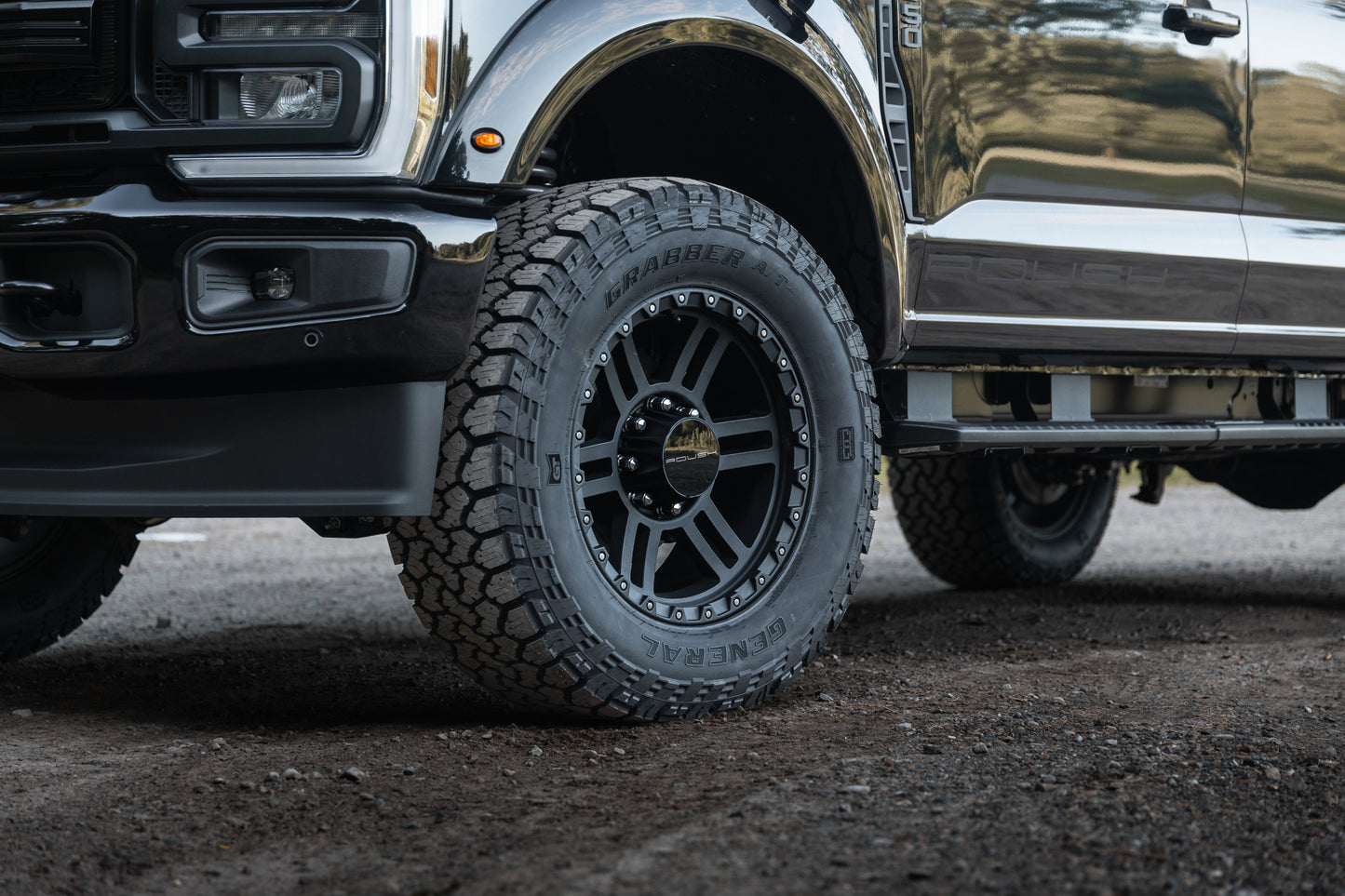 
                  
                    Close-up of a black Roush Super Duty truck tire and wheel parked on a gravel surface, showing the tread and part of the vehicle's front side.
                  
                