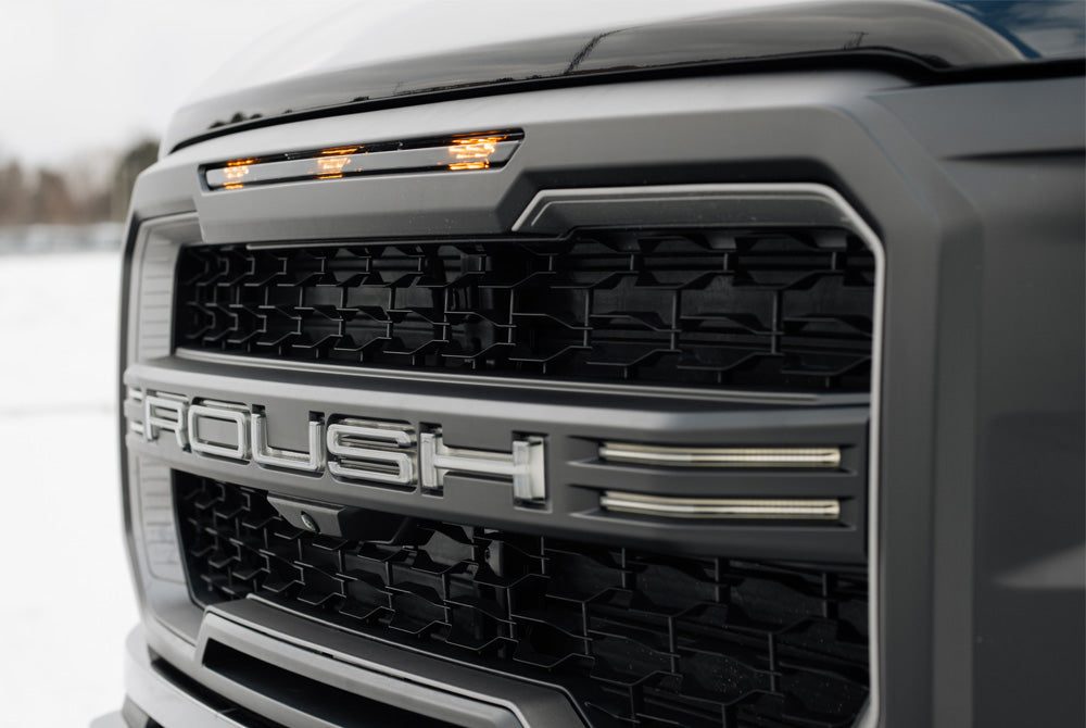 
                  
                    Close-up of a 2025 Roush F-150 grille with bold silver lettering and amber marker lights above.
                  
                