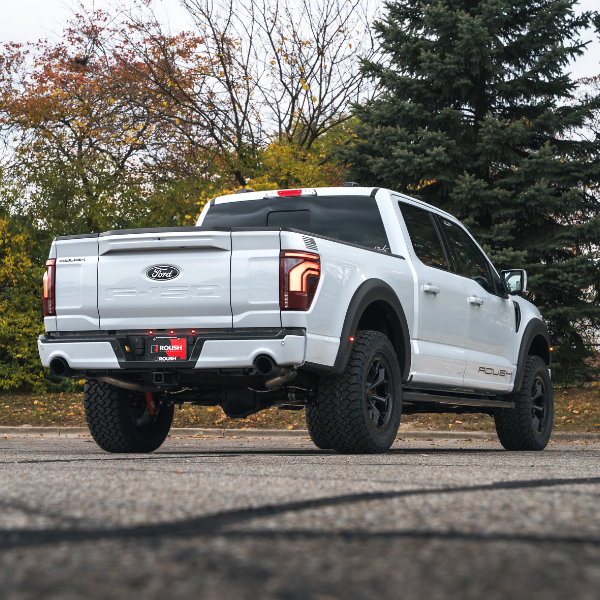 
                  
                    A white Ford pickup truck with black wheels and a sleek rear-exit exhaust system is parked on pavement near trees with fall foliage in the background.
                  
                