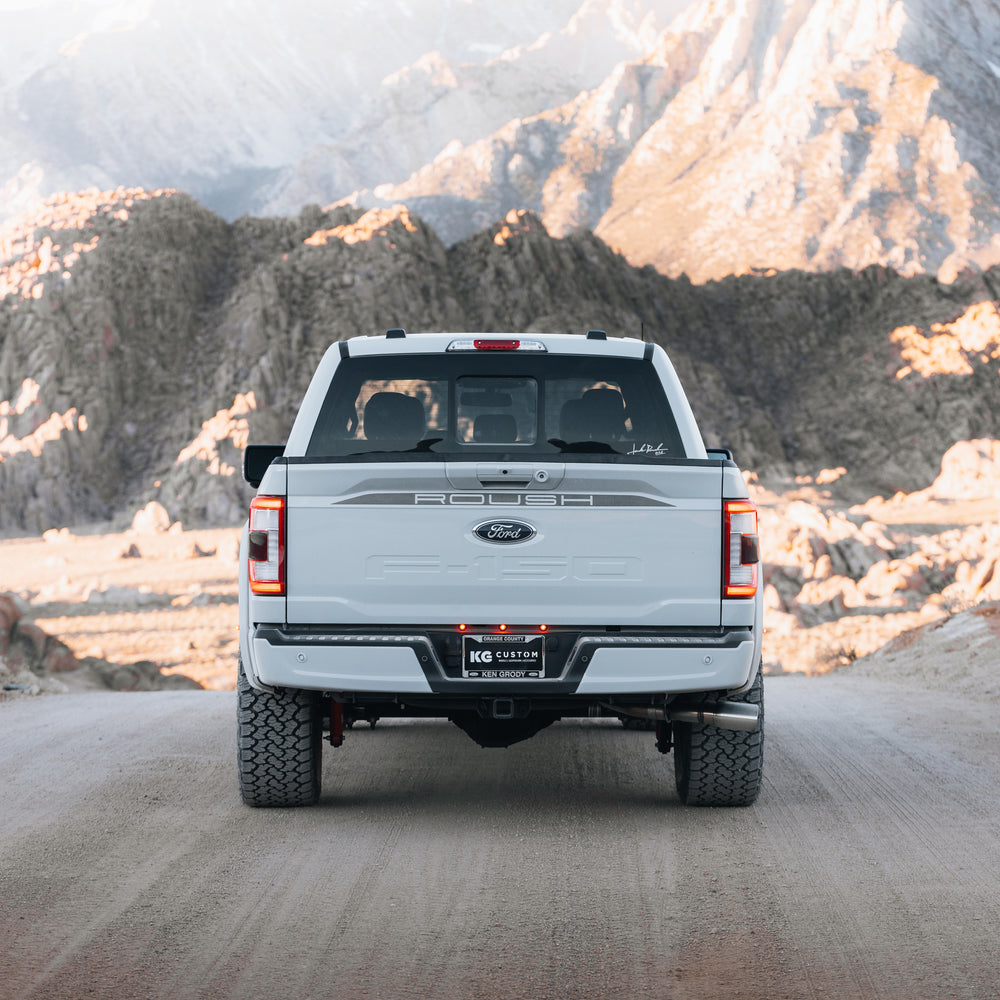 A white 2023 Roush F-150 pickup truck is parked on a dirt road facing away from the camera, showcasing rugged Roush performance enhancements and rocky mountains in the background.