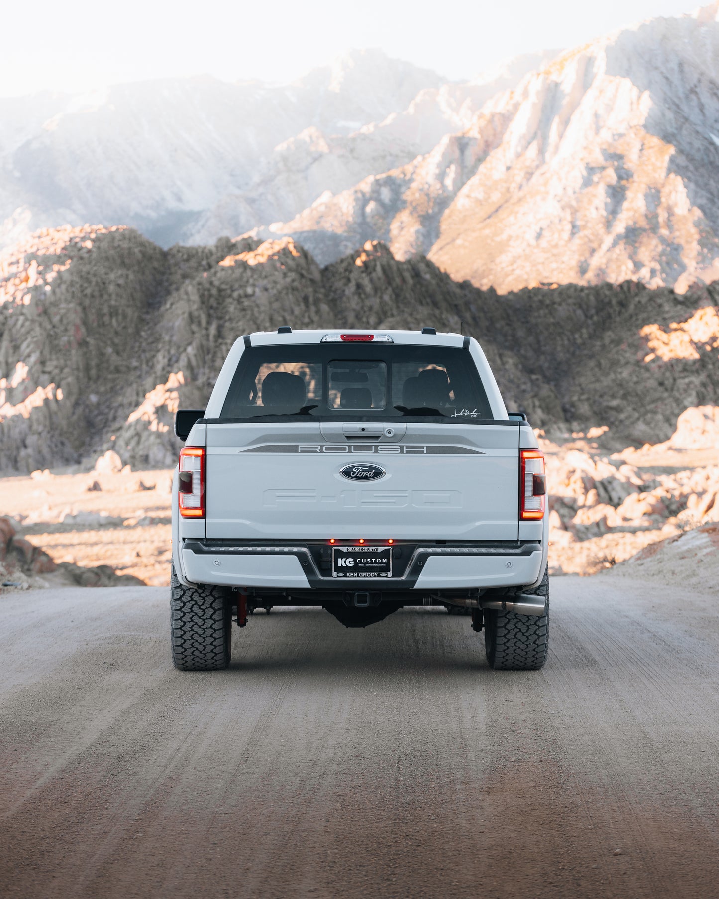 A white 2023 Roush F-150 pickup truck is parked on a dirt road facing away from the camera, showcasing rugged Roush performance enhancements and rocky mountains in the background.