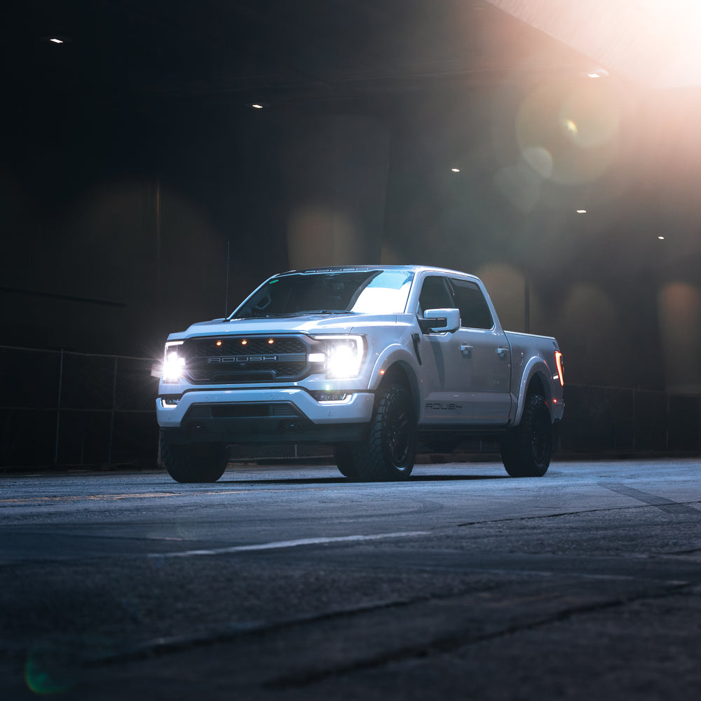 
                  
                    A white 2023 Roush F-150 with headlights on is parked on a dimly lit street under an overpass, showcasing its Roush performance enhancements.
                  
                