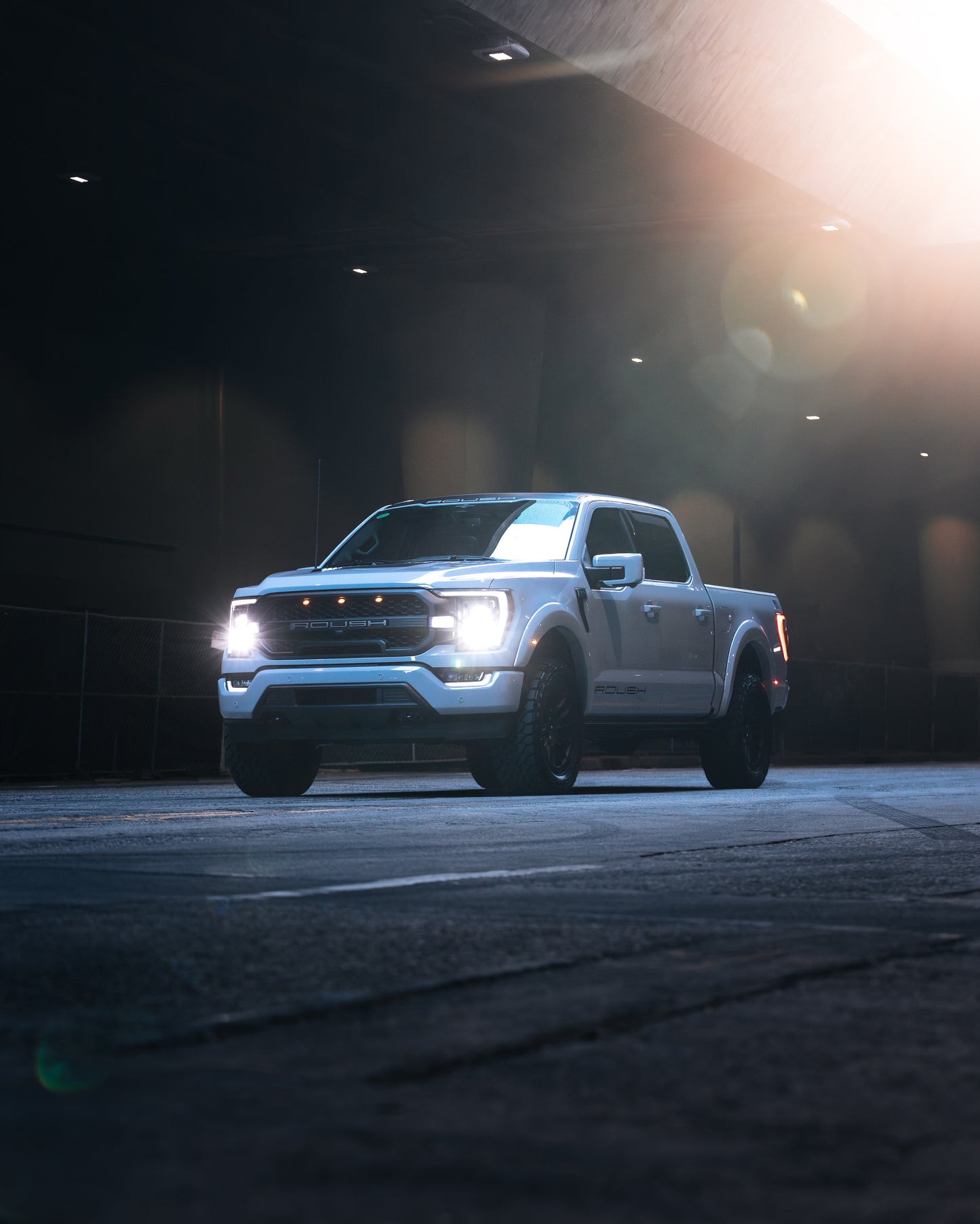 
                  
                    A white 2023 Roush F-150 with headlights on is parked on a dimly lit street under an overpass, showcasing its Roush performance enhancements.
                  
                