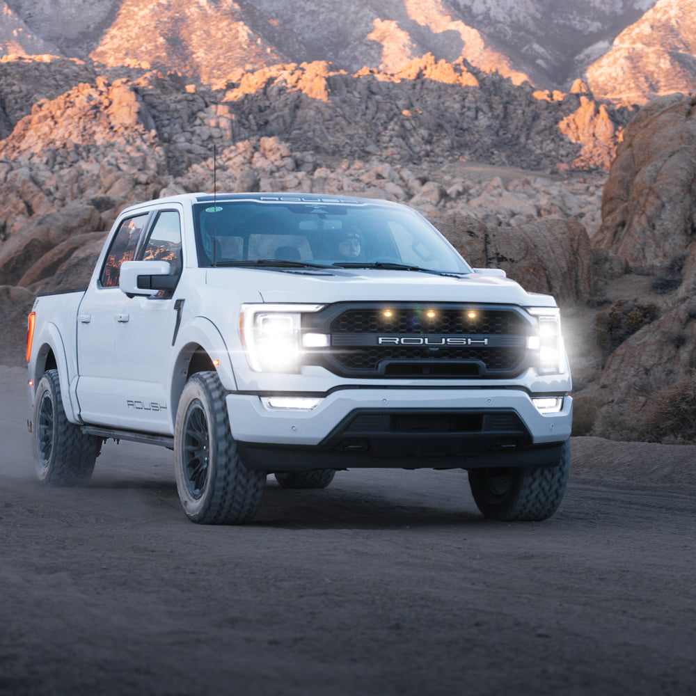 A white 2023 Roush F-150 pickup truck with Roush performance enhancements drives on a dirt road with headlights on, surrounded by rocky terrain and mountains in the background.