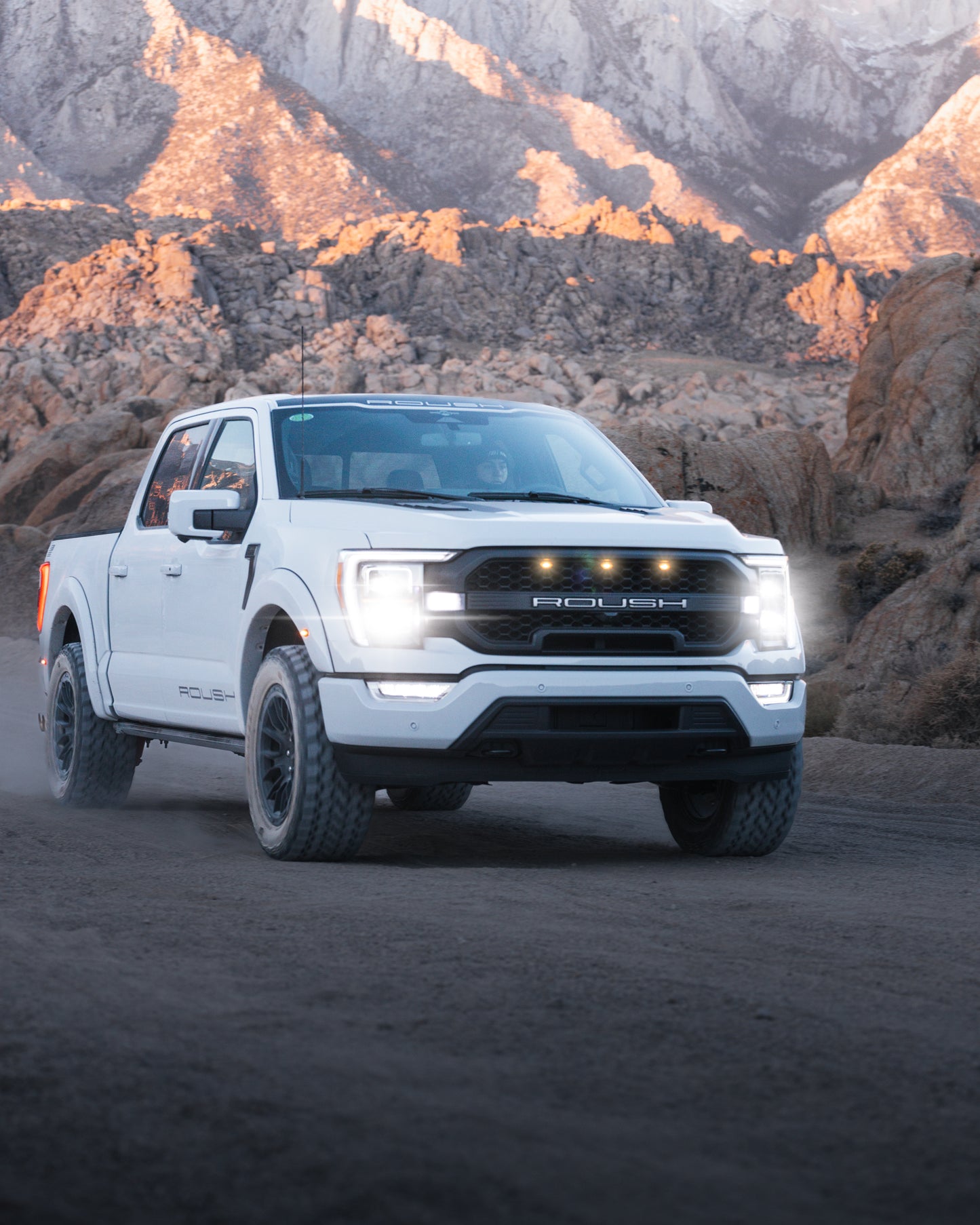 A white 2023 Roush F-150 pickup truck with Roush performance enhancements drives on a dirt road with headlights on, surrounded by rocky terrain and mountains in the background.