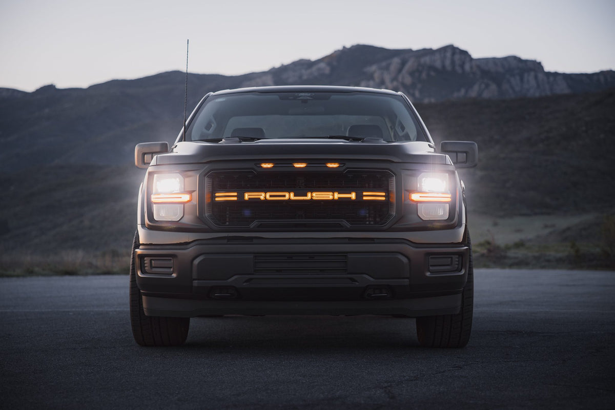 
                  
                    Front view of a dark-colored Roush Nitemare F-150 street performance truck with illuminated grille branding and headlights on.
                  
                