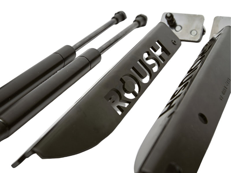 Displayed on a white background are heavy-duty gas struts, specifically designed as Mustang Roush hood struts for the 2005-2014 Mustang, alongside a sleek metal plate with "ROUSH" cut out.