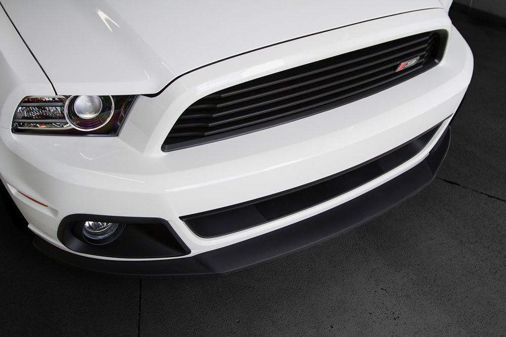 
                  
                    Close-up of the front end of a white Ford Mustang featuring its headlight, bumper, and distinctive Roush Front Grille on a dark concrete surface.
                  
                