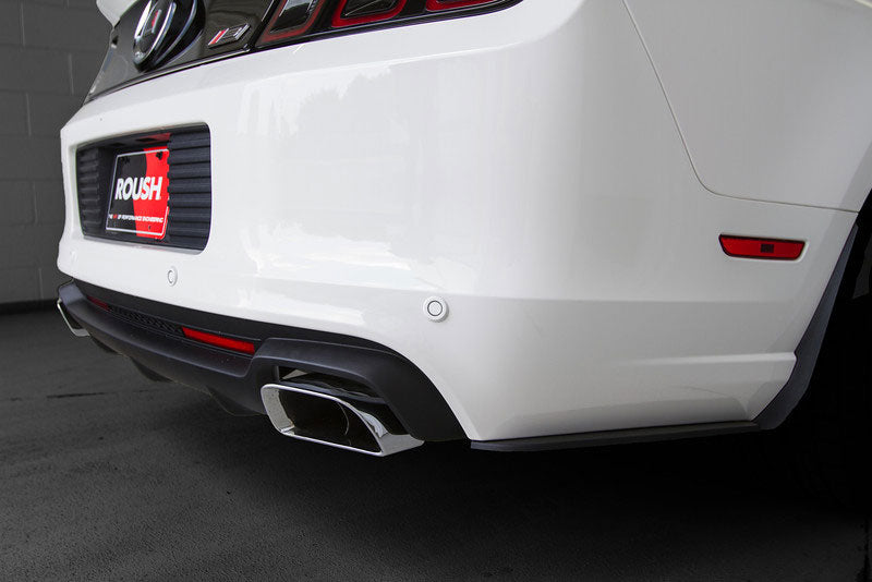 
                  
                    Close-up of the rear bumper and dual exhaust of a white car with a ROUSH license plate frame, featuring a 2013-2014 Mustang Accessories Roush Splitter Kit, parked indoors.
                  
                