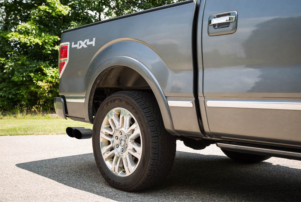 Close-up of the rear side and wheel of a gray Ford F-150 with "4X4" marking and a Roush Performance Exhaust, parked on a paved road near green trees.