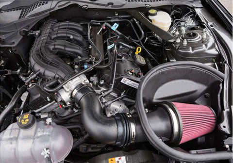 A car engine bay showcasing various components, including a Roush V6 Air Kit with a pink filter, a coolant reservoir, and black engine parts designed for improved engine performance in 2015-2017 Mustangs.