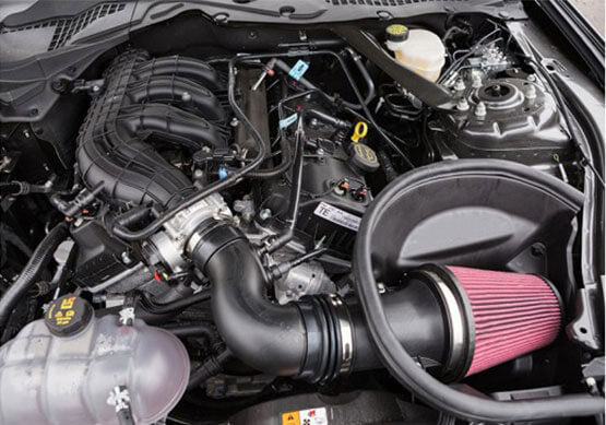 A car engine bay showcasing various components, including a Roush V6 Air Kit with a pink filter, a coolant reservoir, and black engine parts designed for improved engine performance in 2015-2017 Mustangs.