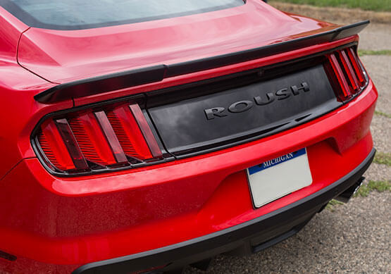 
                  
                    Rear view of a red Roush Mustang, boasting its iconic branding and sleek Mustang rear spoiler. The tail lights illuminate alongside a Michigan license plate, epitomizing the 2015-2023 Mustang's bold style.
                  
                