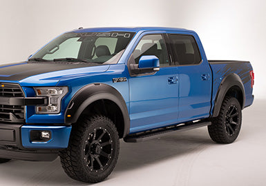 A blue pickup truck with black accents is parked in a well-lit studio setting, showcasing its Roush F-150 Fender Flare Kit. These flares offer exceptional tire coverage and are crafted from UV-protected Dura-Flex 2000 for durability under the spotlight.