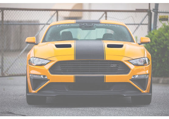 
                  
                    A yellow Mustang GT with black racing stripes and ROUSH Hood Heat Extractors is parked in front of a chain-link fence on a paved surface.
                  
                