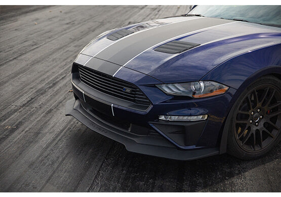 
                  
                    Close-up of the front end of a dark blue Mustang GT with black racing stripes and ROUSH Hood Heat Extractors, parked on a textured asphalt surface.
                  
                