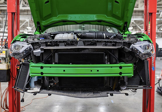 
                  
                    A green car equipped with a ROUSH Stage 3 Max Cooling Upgrade has its front bumper removed and is elevated on a lift in an automotive repair shop, revealing the high-performance Mustang cooling system and internal engine components.
                  
                