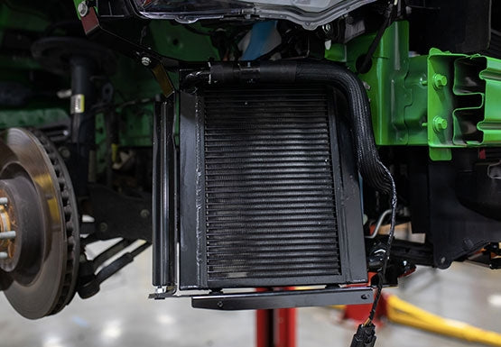 Close-up view of a Mustang cooling system with exposed radiator and cooling components, including the Max Cooling Upgrade; green and black parts surround the area, and a brake disc is visible on the left.