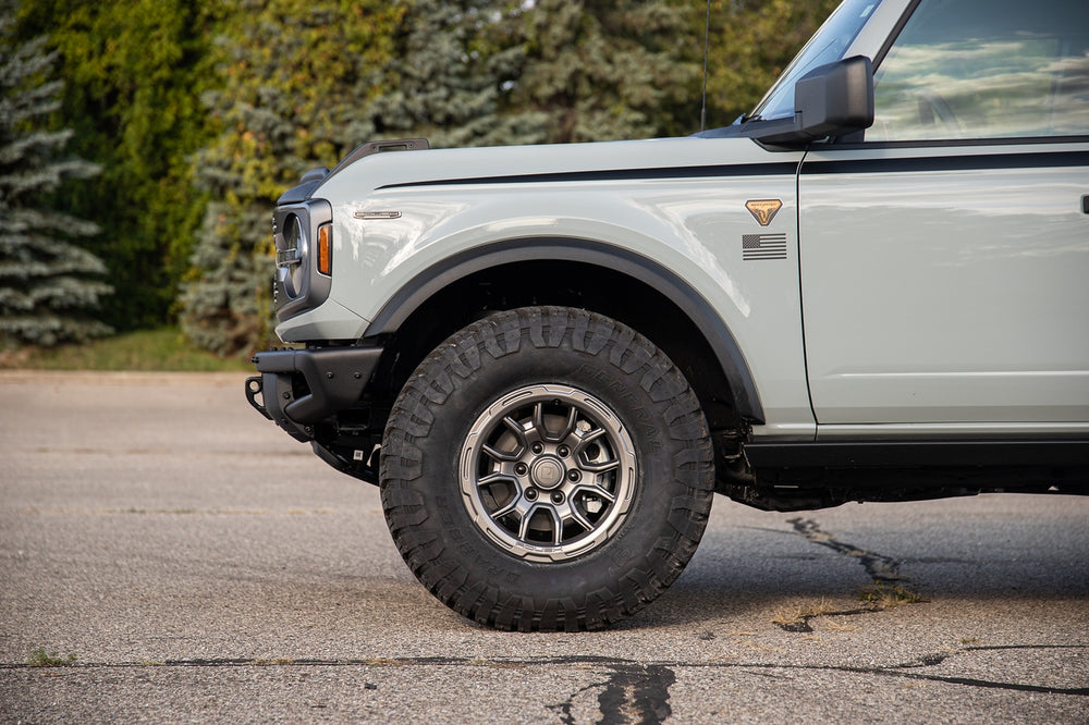 
                  
                    Side view of a parked white SUV focusing on its Roush Performance Products, Inc. 2021-2024 Roush Bronco & Ranger 17-inch Iridium Grey wheel and fender, set against a blurred background of trees.
                  
                