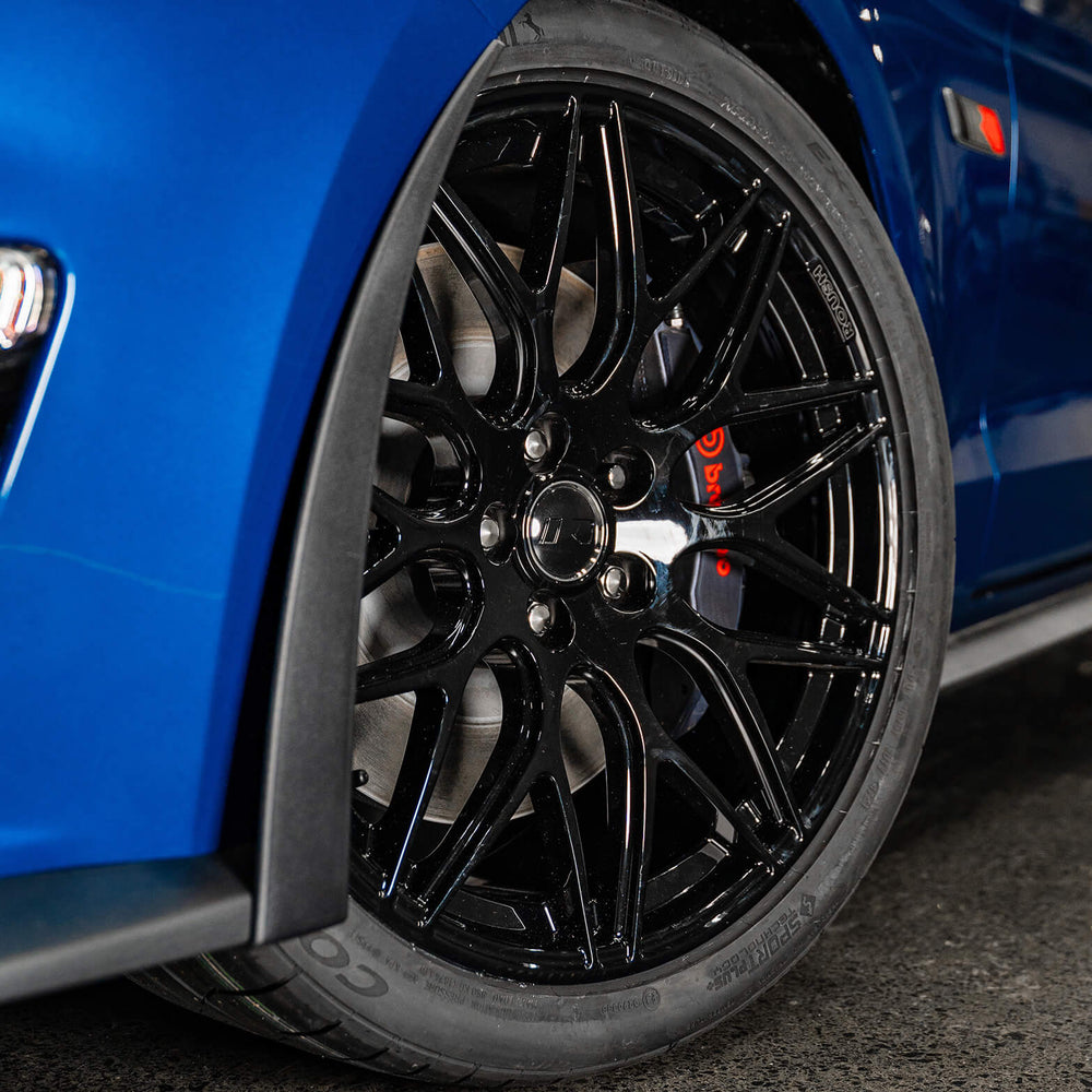 
                  
                    Close-up of a blue ROUSH 450R Mustang's black alloy wheel and red brake caliper, parked on a concrete surface, highlighting its aerodynamic performance.
                  
                