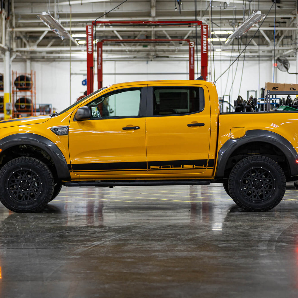 
                  
                    A yellow 2022 Roush Ranger pickup truck, known for its off-road capability, is parked inside an industrial garage, with equipment and machinery visible in the background. The truck features black wheels and decals along the sides.
                  
                