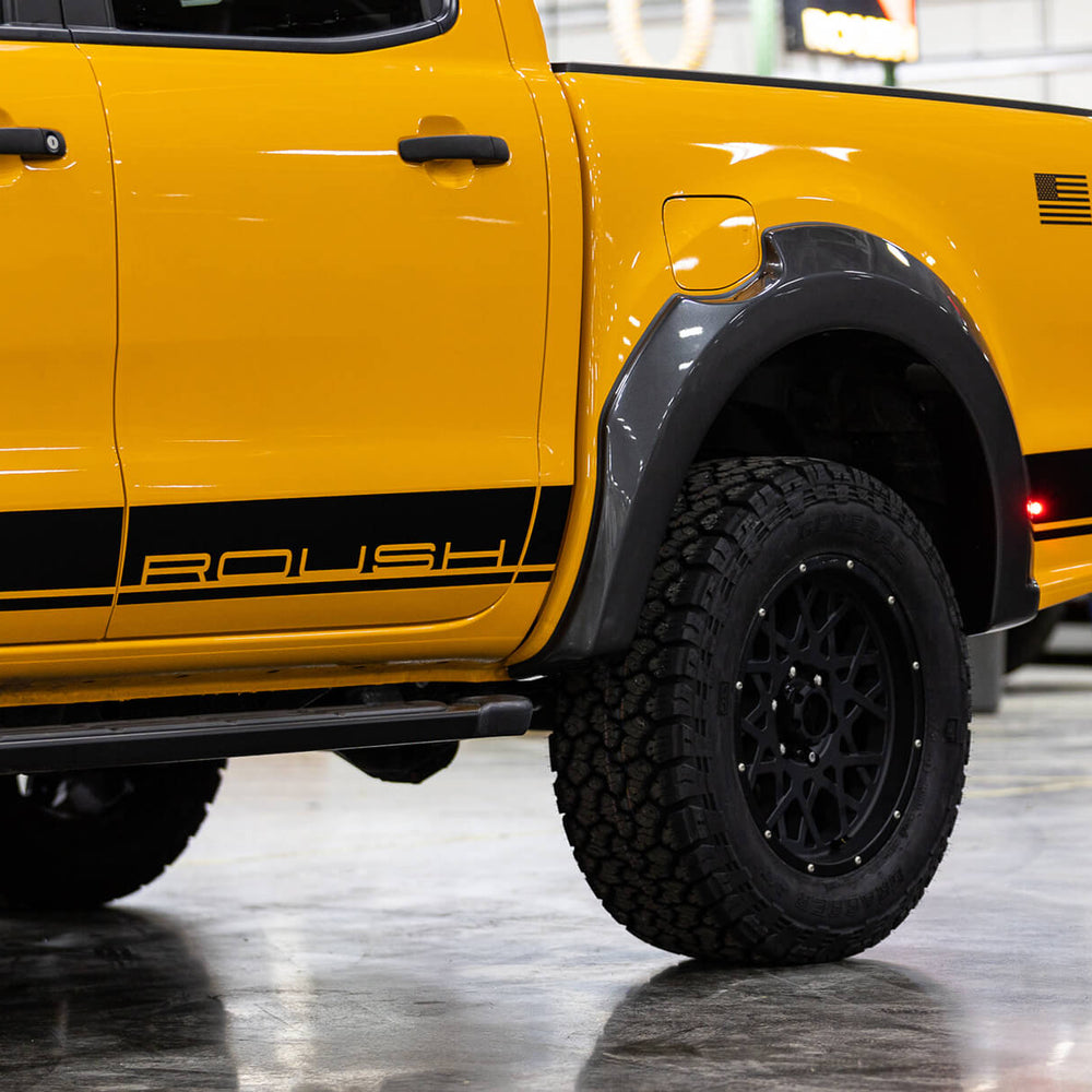 
                  
                    Close-up of a bright yellow 2022 Roush Ranger with black ROUSH branding. The truck, showcasing its impressive off-road capability, is parked in a garage or workshop, with a stack of tires visible in the background.
                  
                