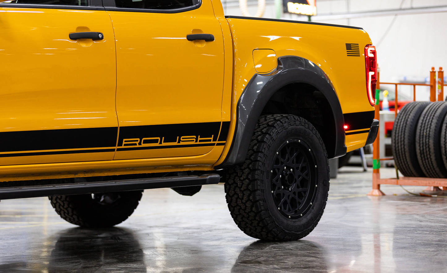 
                  
                    Close-up of a bright yellow 2022 Roush Ranger with black ROUSH branding. The truck, showcasing its impressive off-road capability, is parked in a garage or workshop, with a stack of tires visible in the background.
                  
                