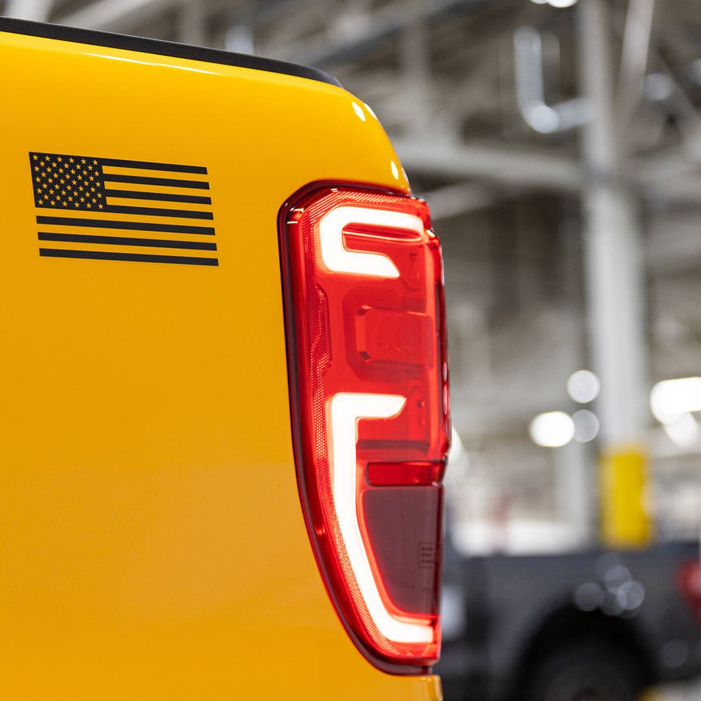 
                  
                    Close-up of the rear section of a yellow 2022 Roush Ranger, featuring an American flag decal and distinct LED taillight with a white and red design. The background displays a blurred industrial setting, hinting at the truck's off-road capability.
                  
                