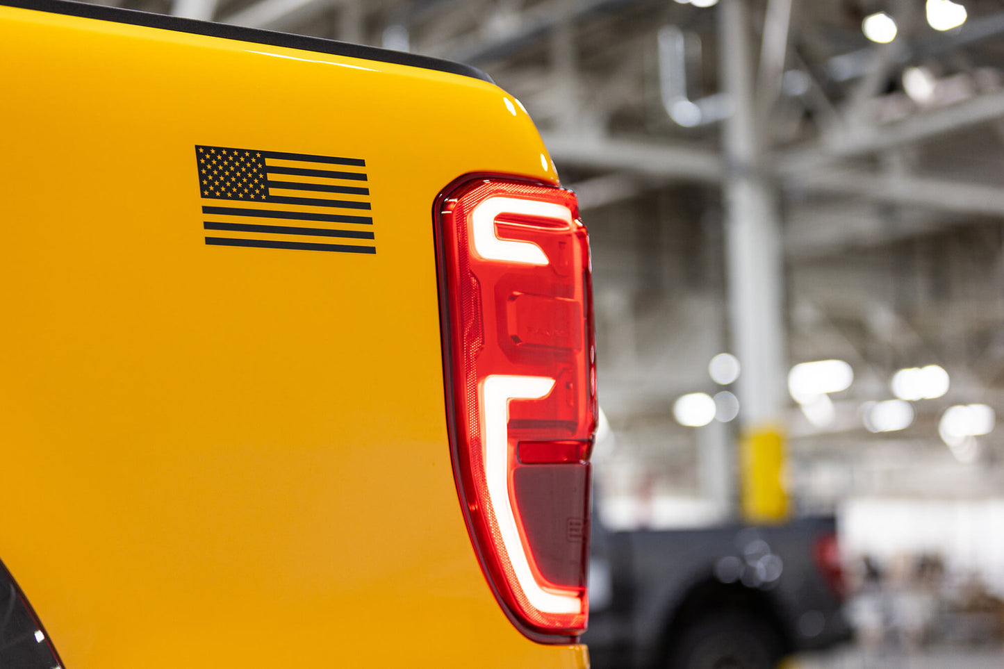
                  
                    Close-up of the rear section of a yellow 2022 Roush Ranger, featuring an American flag decal and distinct LED taillight with a white and red design. The background displays a blurred industrial setting, hinting at the truck's off-road capability.
                  
                