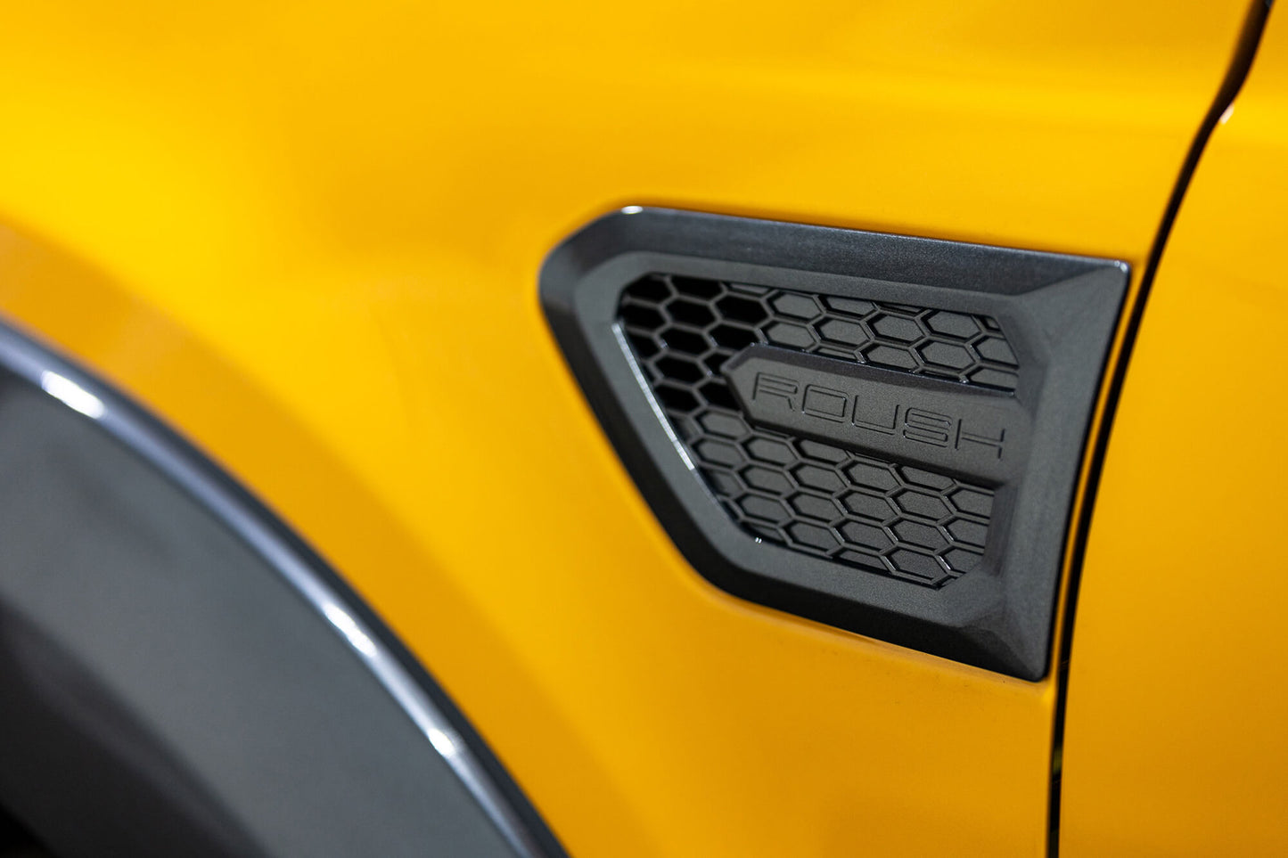 
                  
                    Close-up of a yellow 2022 Roush Ranger with a black, hexagon-patterned vent labeled "ROUSH" on the side, showcasing its off-road capability.
                  
                
