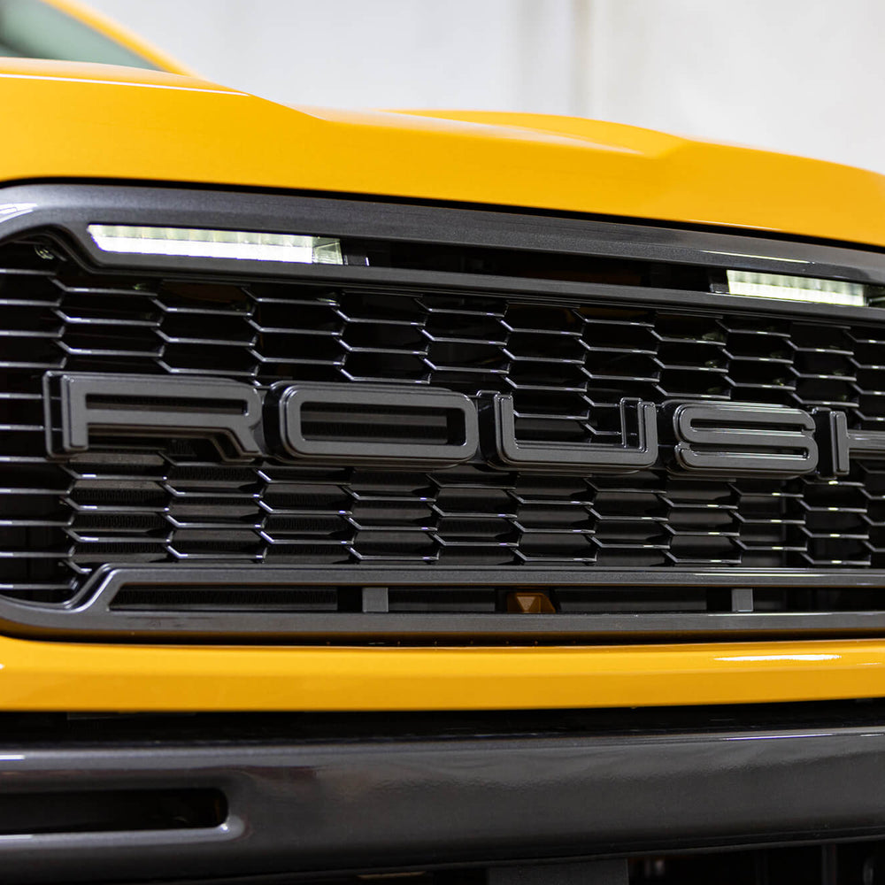 
                  
                    Close-up of the front grille of a yellow 2022 Roush Ranger with the word "ROUSH" prominently displayed in the center, showcasing its impressive Roush Performance.
                  
                