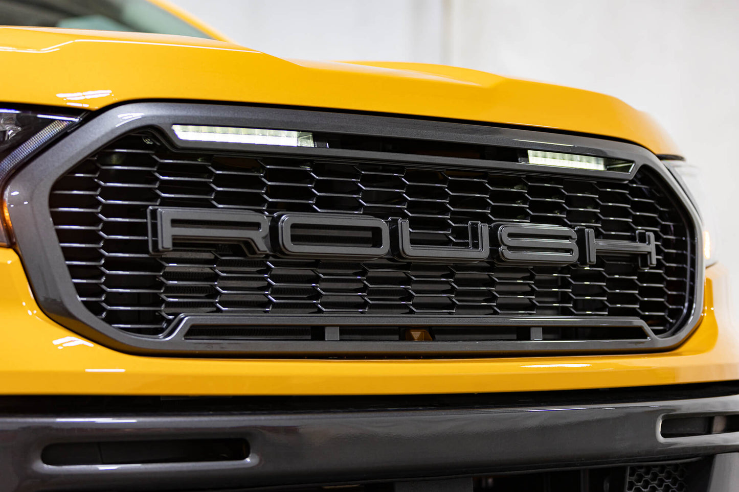 
                  
                    Close-up of the front grille of a yellow 2022 Roush Ranger with the word "ROUSH" prominently displayed in the center, showcasing its impressive Roush Performance.
                  
                