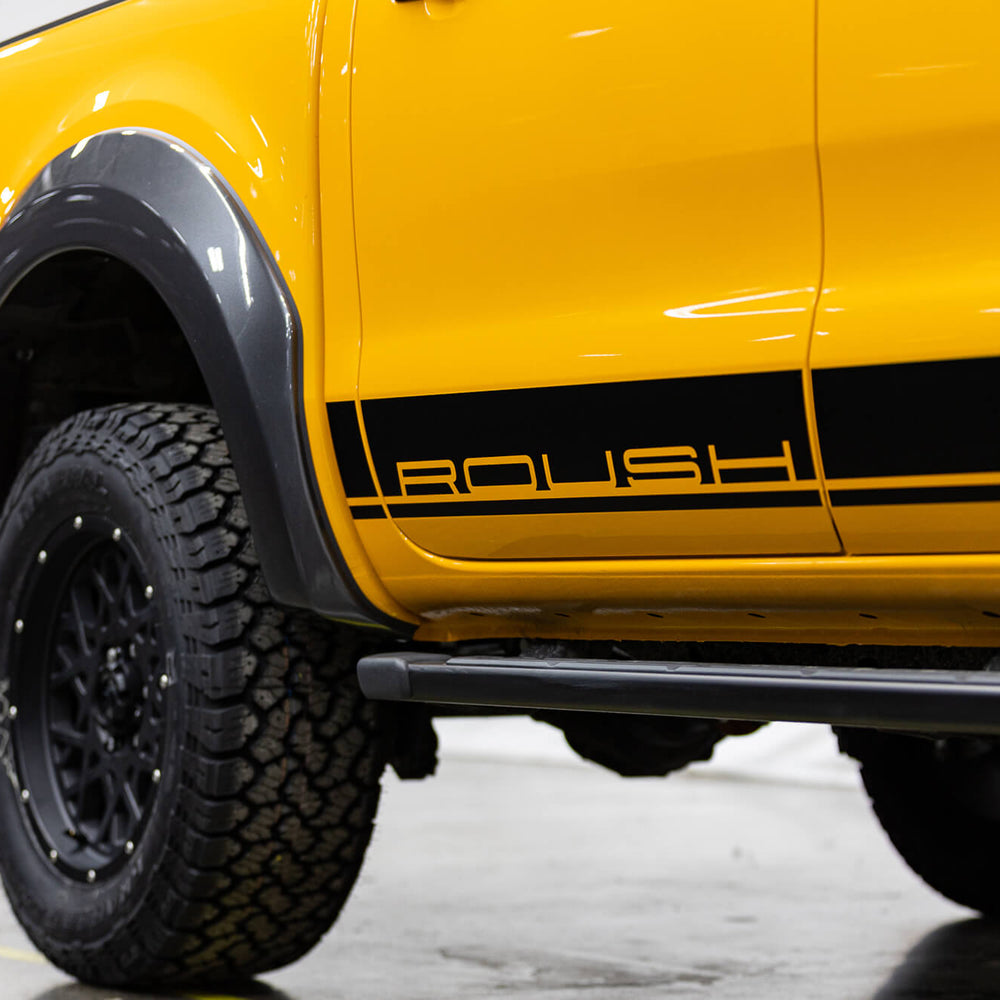 
                  
                    Photo of a 2022 Roush Ranger's rear side with black text "ROUSH" above a black stripe near the bottom, black fender flares, and large black off-road tires, highlighting its off-road capability and parked indoors.
                  
                
