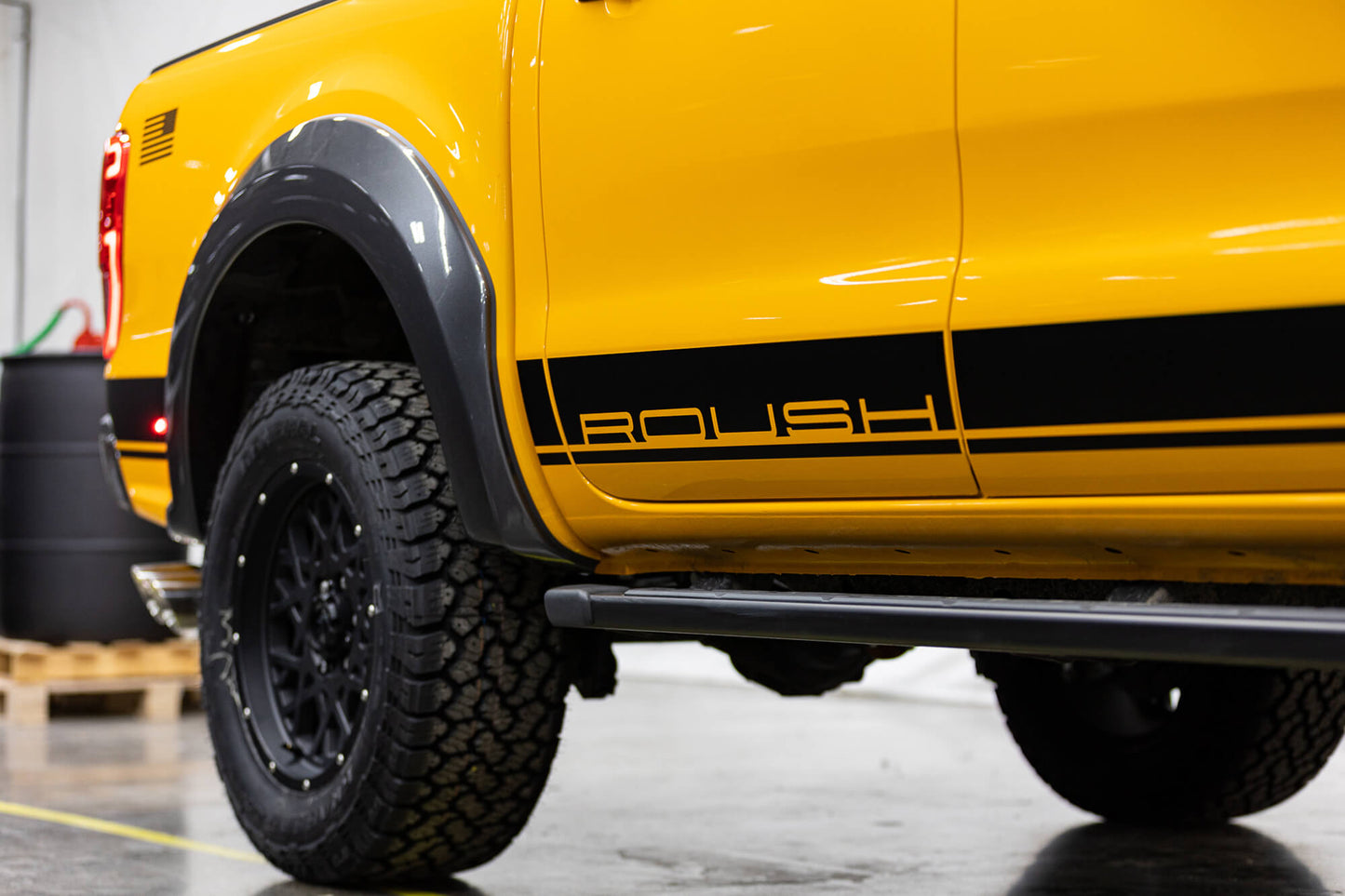
                  
                    Photo of a 2022 Roush Ranger's rear side with black text "ROUSH" above a black stripe near the bottom, black fender flares, and large black off-road tires, highlighting its off-road capability and parked indoors.
                  
                