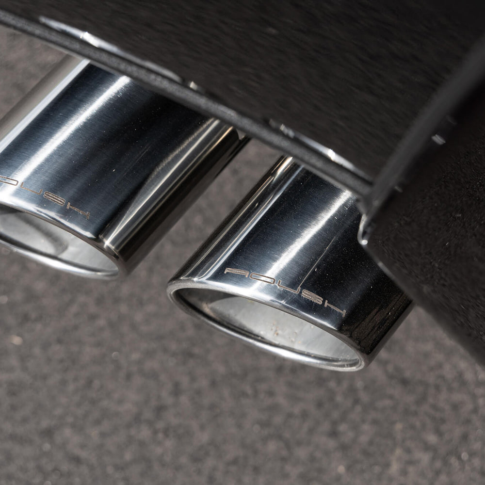 
                  
                    Close-up view of dual chrome exhaust tips on a black vehicle, marked with the Porsche logo, set against a gray pavement background, adjacent to the ROUSH High-Flow Front Grille.
                  
                