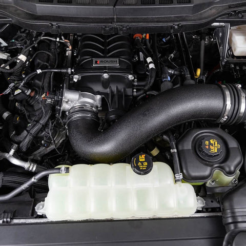 A close-up view of a car engine compartment showcasing various components, including the Roush Performance F-150 Supercharger Kit, air intake, battery, coolant reservoir, and other engine parts that contribute to its impressive 705 horsepower.