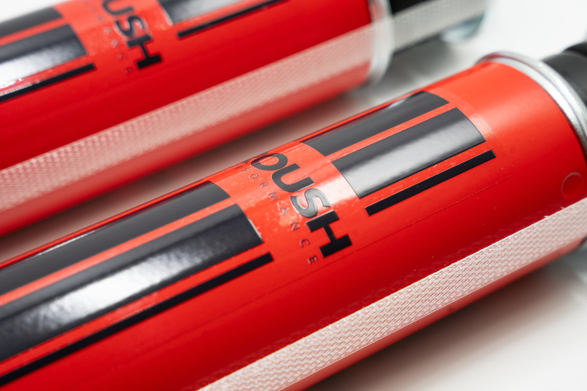 
                  
                    Close-up of two red cylindrical objects with black and white details and the word “ROUSH” printed on the surface, showcasing components from a Roush Ram 1500 suspension performance suspension system.
                  
                