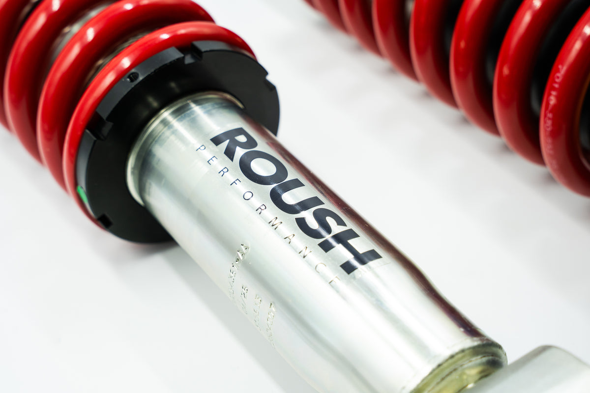 
                  
                    Close-up of a silver ROUSH Performance car shock absorber with red coil springs, showcasing the advanced performance suspension system, against a white background.
                  
                