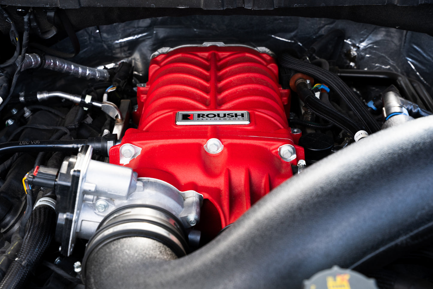 
                  
                    Close-up of a red Roush supercharger kit installed in a vehicle engine bay, surrounded by black hoses and metal components, designed to boost your F-150's power up to 705 horsepower.
                  
                