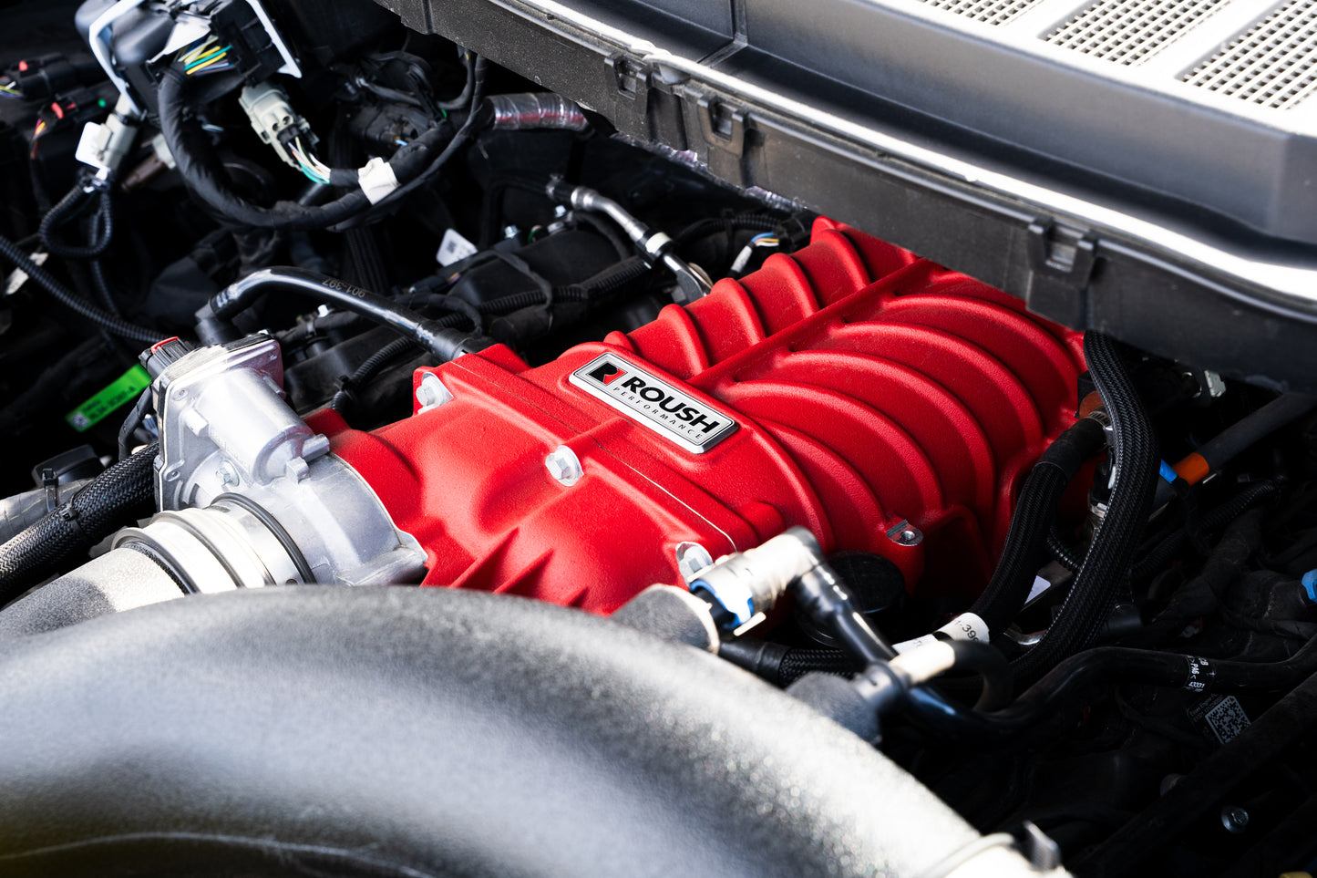 
                  
                    Close-up of a red Roush supercharger kit installed on a car engine, surrounded by various black hoses and engine components, unleashing up to 705 horsepower.
                  
                
