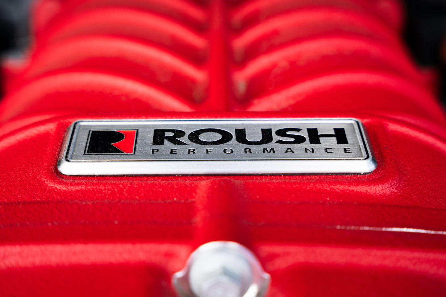 
                  
                    Close-up of a red engine cover featuring a silver and black "Roush Performance" emblem, highlighting the power behind the 705 horsepower Roush supercharger kit.
                  
                