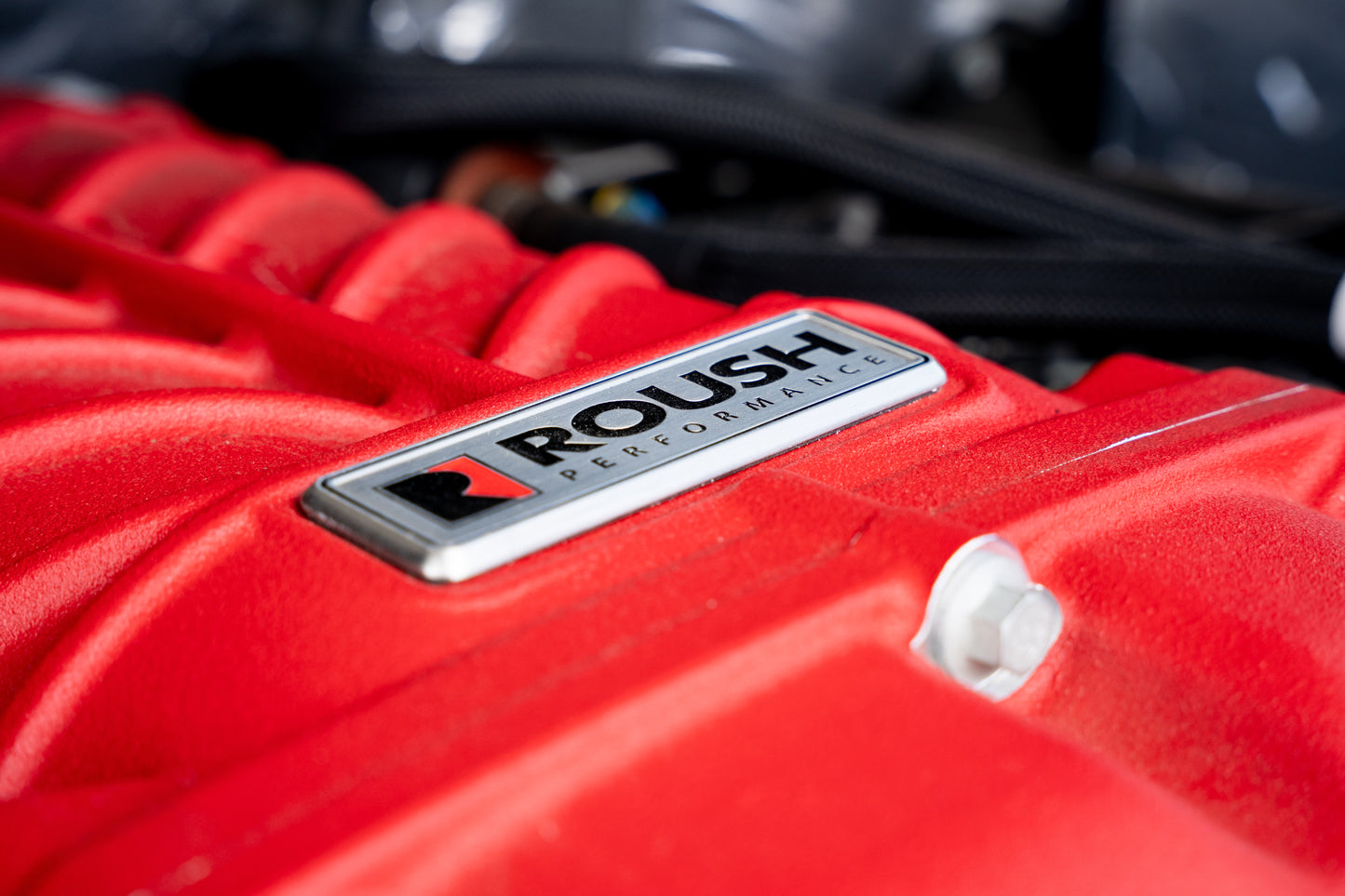 
                  
                    Close-up of a red engine cover featuring a "ROUSH Performance" badge, textured metal, and bolts—highlighting the power of a Roush supercharger kit that drives up to 705 horsepower.
                  
                