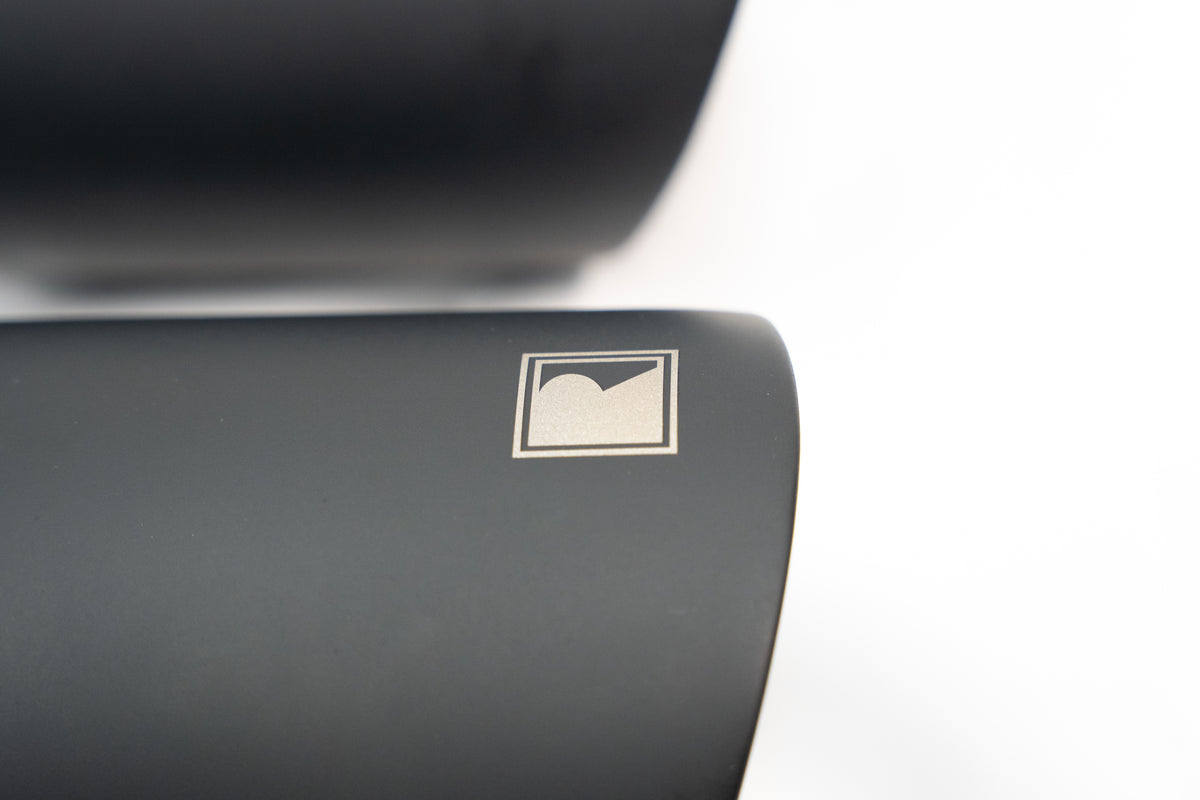 
                  
                    Close-up of a matte black object, reminiscent of a sleek black tip exhaust, featuring a small, engraved geometric logo with a circle and a sharp corner on a white background.
                  
                
