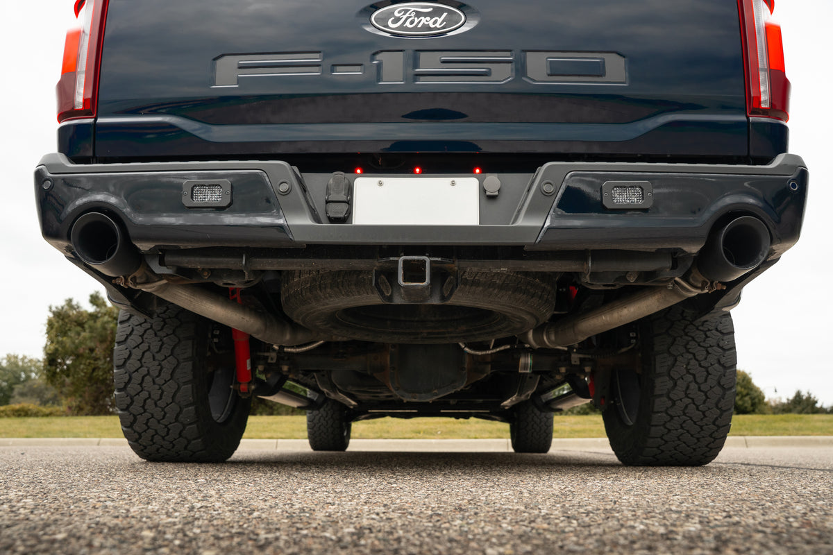 
                  
                    Rear view of a Ford F-150 truck, focusing on the bumper, Roush F-150 cat-back exhaust with black tip rear-exit exhaust, spare tire, and undercarriage, parked on a paved surface.
                  
                