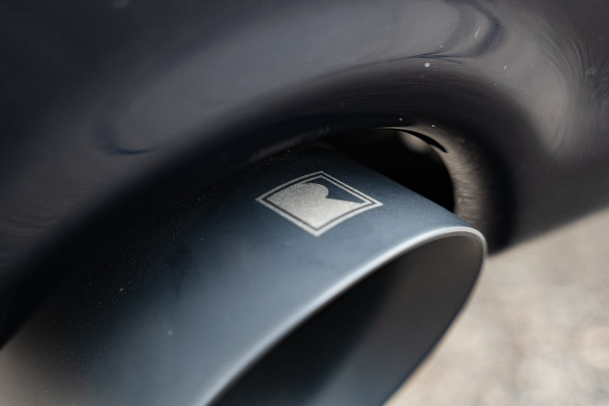 
                  
                    Close-up of a car’s exhaust pipe with a rectangular logo featuring a stylized “R” on its surface, showcasing the premium design of the Roush F-150 exhaust.
                  
                