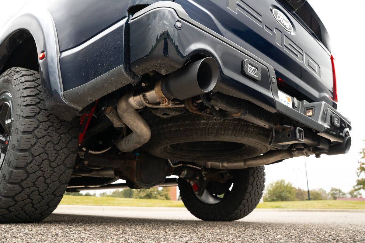 
                  
                    Low-angle view of the rear underside of a Ford F-150 truck, showcasing the performance exhaust system with a black tip rear-exit exhaust, along with the spare tire and part of the rear bumper.
                  
                