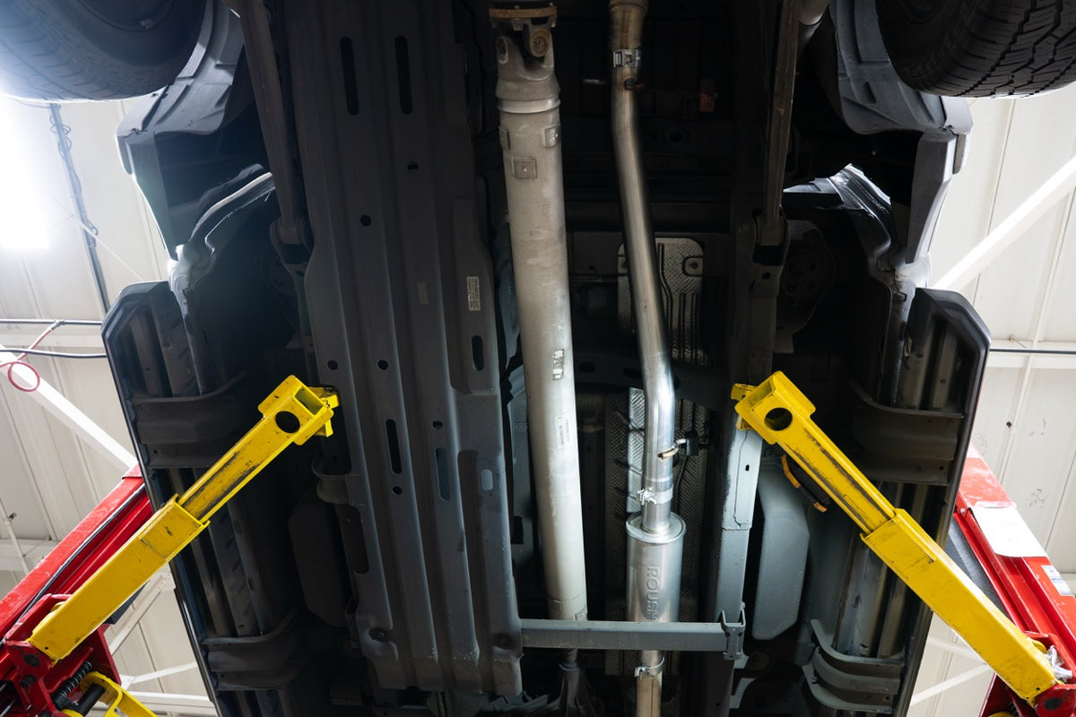 
                  
                    Undercarriage of a Ram 1500 5.7L HEMI raised on a yellow hydraulic lift, showing the driveshaft and a Roush performance exhaust system.
                  
                