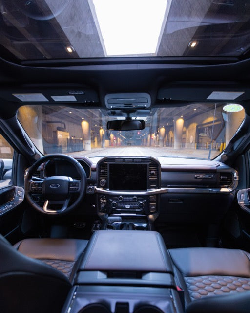 
                  
                    View from the interior of a 2023 Roush F-150 showcasing Roush performance enhancements, with the dashboard, steering wheel, and large sunroof framing an urban street scene through the windows.
                  
                