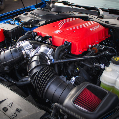 Close-up of a car engine with a red cover featuring the Roush logo. The supercharger kit powers this beast to an impressive 740 horsepower. Black hoses and various engine components are visible, with the bottom of the hood showcasing "Mustang" text partially seen, hinting at its Roush S650 Mustang heritage.